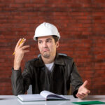 front view male engineer sitting his working place white helmet document business corporate property job builder contractor plan